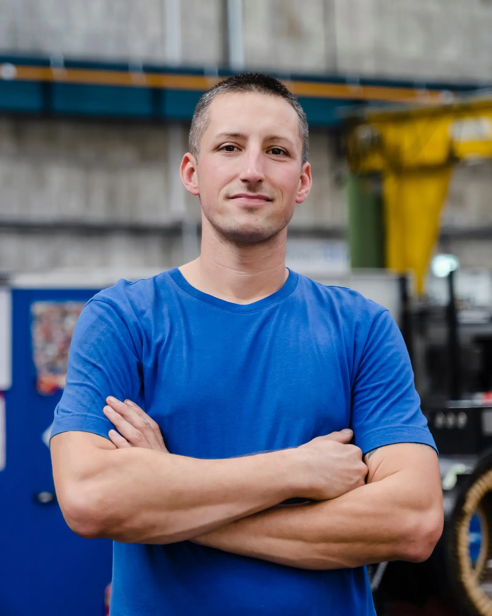Ein Fabrikarbeiter in einem blauen Shirt steht mit verschränkten Armen in der Werkhalle.