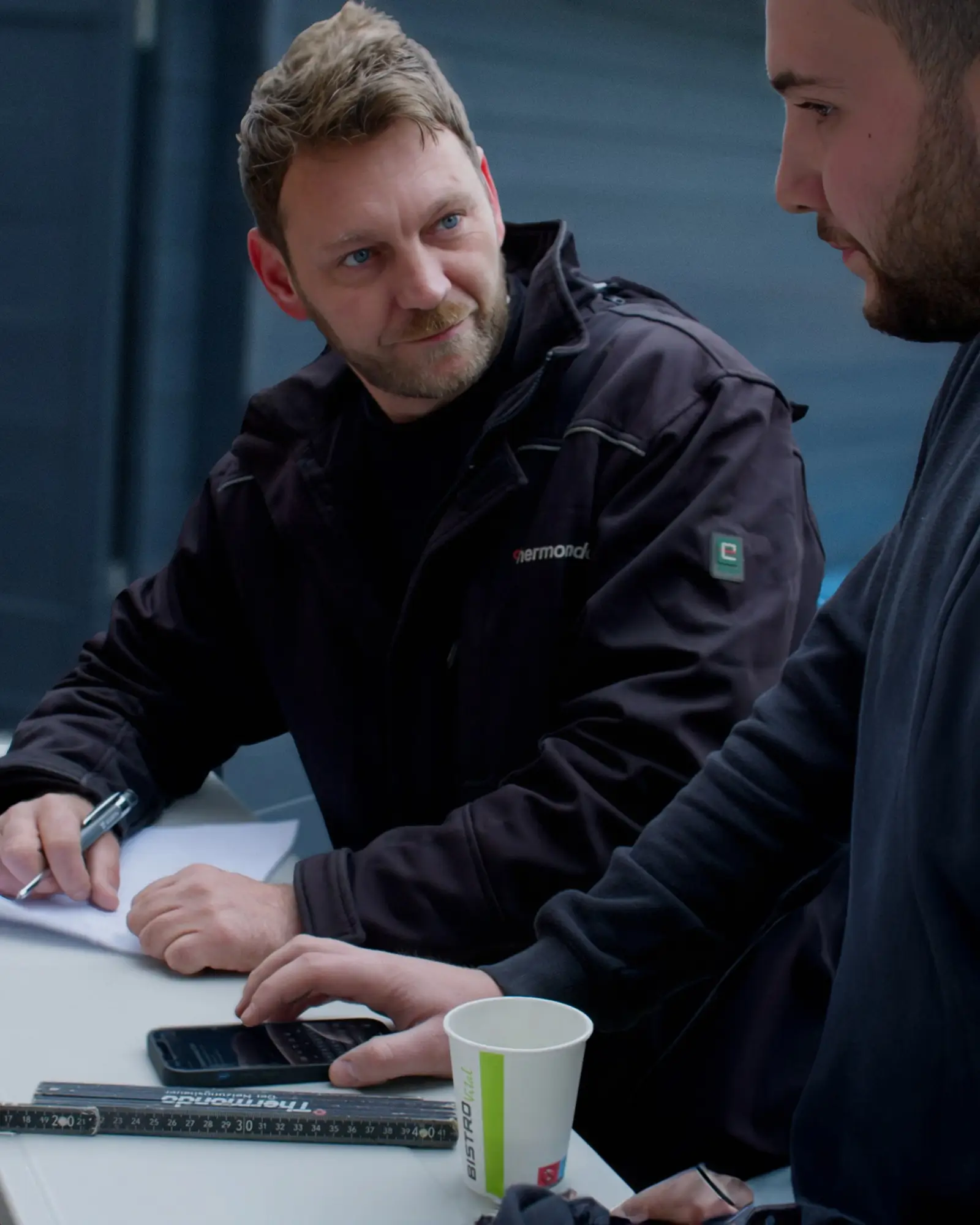 Zwei Kollegen besprechen Notizen und einen Laptop an einem Arbeitsplatz im Freien.